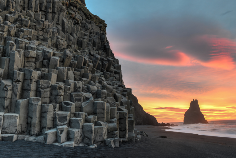 Picture of Reynisfjara black sand beach in Iceland with basalt rock formations and orange sunset over the ocean in the background