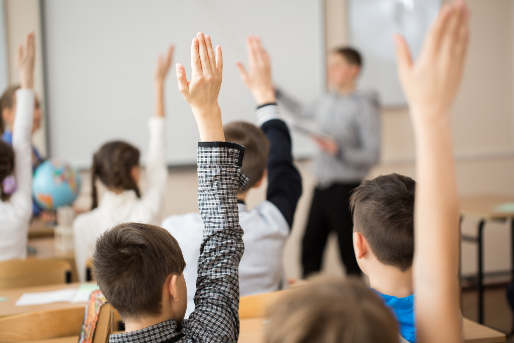 picture of students in a classroom raising their hands with a teacher in front of a white board
